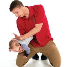Homme avec un chandail rouge qui tient un bébé d'une main, et qui tape entre ses omoplates de l'autre main