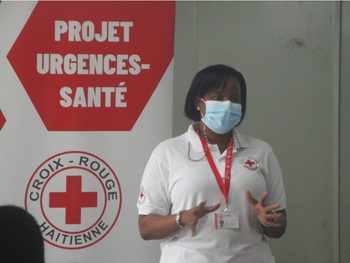 Une femme portant un masque et un chandail blanc avec le logo de la Croix-Rouge  se tient debout devant un panneau blanc