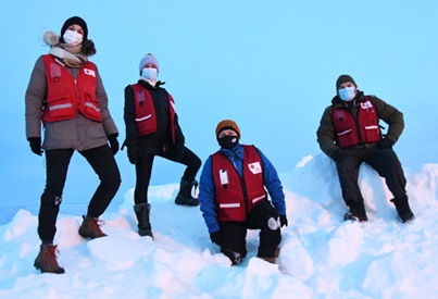 Quatre bénévoles de la Croix-Rouge canadienne, portant des gilets rouges par-dessus leurs manteaux d'hiver, escaladent une montagne de neige. Ils portent tous un masque médical.