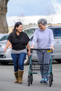 An elderly woman using a walker with a cart walks alongside another woman in a parking lot.