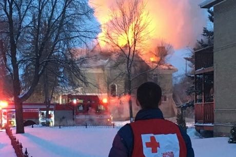 Un bénvole de la Croix-Rouge se tient debout de dos devant une maison en flammes durant l'hiver.