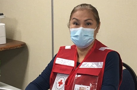 Une femme, portant une veste rouge de la Croix-Rouge et un masque, assise à un bureau