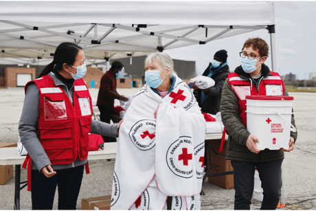 Two Red Cross members walking with an older woman between them who is wrapped in a Red Cross blanket