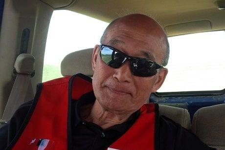 Un homme assis dans une voiture avec des lunettes de soleil noires et une veste de la Croix-Rouge sourit à la caméra.