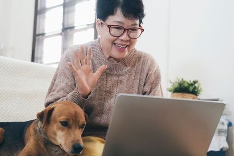 Une femme d'origine asiatique avec un chandail beige et des lunettes rouges sourit et envoie la main à la caméra sur son ordinateur avec son chien à côté d'elle.
