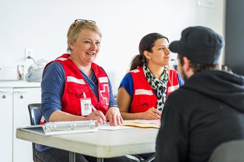 Deux personnes portant un dossard de la Croix-Rouge parlent à une personne assise devant elles. Les travailleurs ont un stylo en main et des formulaires sont sur la table.