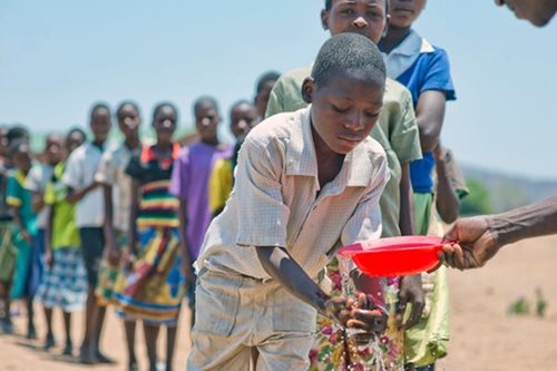 Une file d’enfants dans un paysage sec et ensoleillé. L’enfant à l’avant de la file se lave les mains dans un bol rouge rempli d’eau.