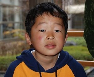 Un petit garçon japonais avec un chandail orange et bleu regarde la caméra.