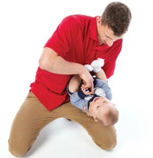 Homme accroupi, avec un chandail rouge, qui tient un bébé sur sa jambe, avec une main sous sa tête, et l'autre main qui lui touche le cou