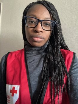 Une femme à la peau noire avec des lunettes noires et une veste de la Croix-Rouge sourit à la caméra.