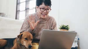 Une femme d'origine asiatique avec un chandail beige et des lunettes rouges sourit et envoie la main à la caméra sur son ordinateur avec son chien à côté d'elle.