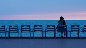 Une femme assise de dos devant l'océan au coucher de soleil avec plusieurs chaises vides à côté d'elle.