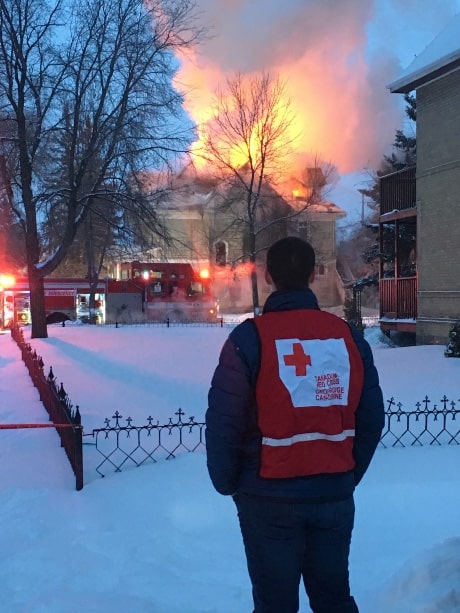Un bénévole de la Croix-Rouge se tient debout de dos devant une maison en flammes durant l'hiver.