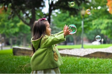 Petite fille d'origine asiatique avec un manteau vert qui court pour attraper des bulles.
