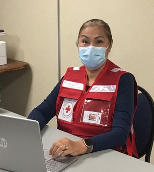 Une femme, portant une veste rouge de la Croix-Rouge et un masque, assise à un bureau