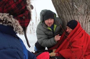 Un homme assis par terre, avec une couverture rouge sur les épaules, souffrant d'hypothermie, avec un autre homme près de lui qui l'aide