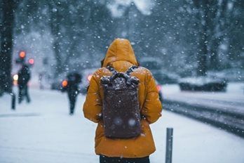 Une personne, portant un manteau d'hiver jaune et un sac à dos noir, qui marche sur un trottoir lors d'une tempête de neige