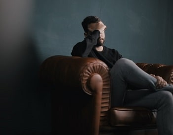 Un homme se tient le front, assis sur un divan dans un décors sombre