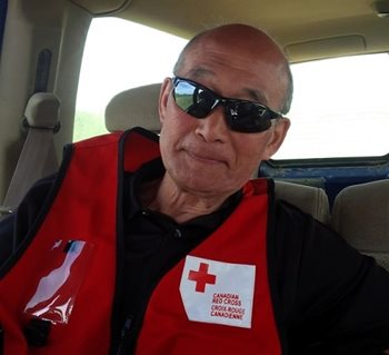 Un homme assis dans une voiture avec des lunettes de soleil noires et une veste de la Croix-Rouge sourit à la caméra.