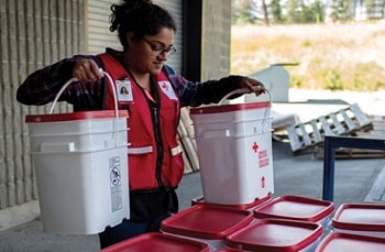 Une femme arborant un dossard de la Croix-Rouge soulève deux seaux blancs au couvercle rouge qui contiennent des trousses d’urgence.