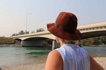 Une femme portant un chapeau et une camisole en jeans de dos devant une rivière et un pont