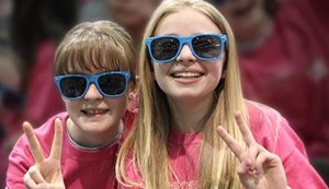 Deux jeunes filles vêtues de chandails rose et de lunettes bleus font un signe de paix en souriant