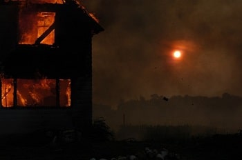 Une maison brûle avec en arrière-plan un champ et la lune qui brille à travers la fumée.