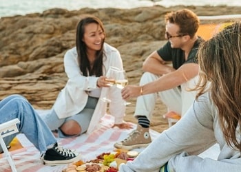 Deux femmes et un homme sont assis sur une couverture blanche à la plage. Ils discutent en riant autour d’un pique-nique. Un goéland les guette en arrière-plan.