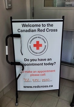 Devant l’entrée du bureau de la Croix Rouge à Dartmouth, en Nouvelle Écosse, une canne noire à poignée désaxée est appuyée à la verticale devant une pancarte blanche de la Croix Rouge canadienne. Le logo de la Croix-Rouge apparaît dans le haut de la pancarte. Le texte suivant y est inscrit en anglais, en caractères noirs : « Welcome to the Canadian Red Cross. Do you have an appointment today? ». Cela signifie : « Bienvenue à la Croix Rouge canadienne, avez-vous un rendez-vous aujourd’hui? ».