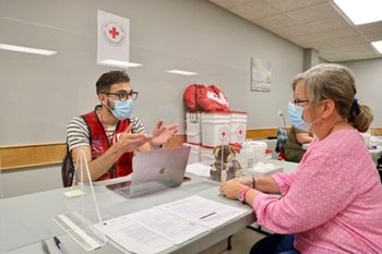 Karar, vêtu d'un masque et d'un gilet de la Croix-Rouge, assis derrière une feuille de plexiglas, avec un résident de la Colombie-Britannique assis en face de lui.