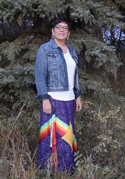 Une femme autochtone debout devant de grands arbres, portant une jupe et une veste en jean.