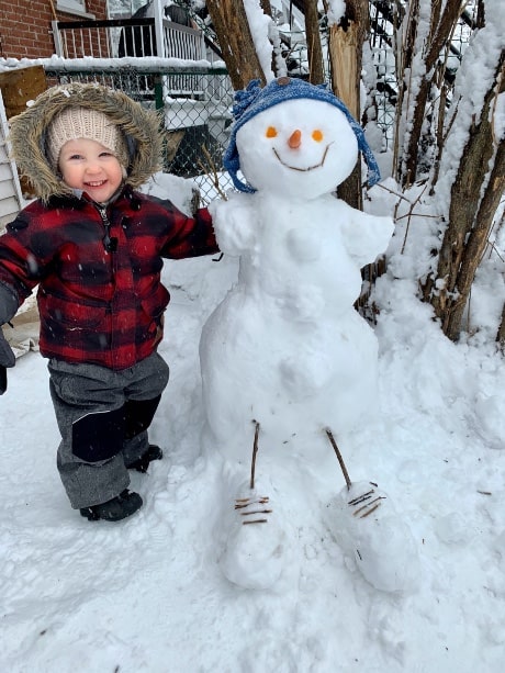 Enfant dans un ensemble d'hiver rouge et noir, avec une tuque beige, se tenant à côté d'un bonhomme de neige