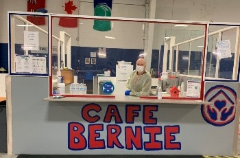 Un responsable de la Croix-Rouge est accoté sur un comptoir nommé Café Bernie avec un masque et une blouse de protection contre la COVID-19.