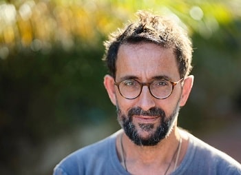 Photo prise en plein air d’un homme aux cheveux foncés, qui a une barbe noire et grise et la peau bronzée et sourit légèrement. Il porte un t-shirt bleu et des lunettes.