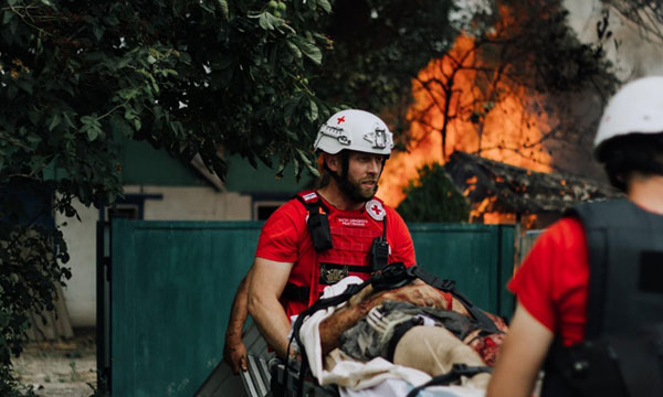 Deux médecins de la Croix-Rouge transportent une personne blessée sur une civière, alors qu'un incendie ravage un arbre et un bâtiment en arrière-plan.