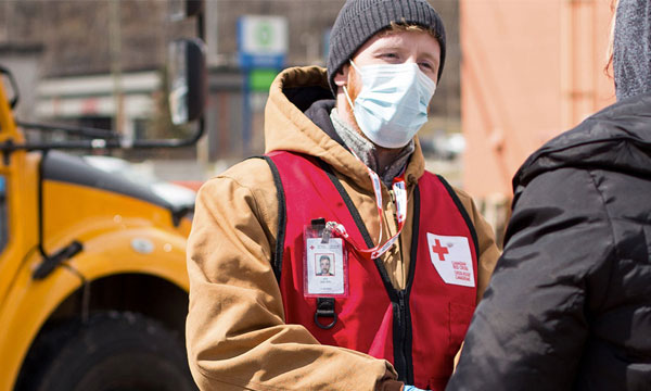 Un bénévole de la Croix-Rouge canadienne porte un masque médical et se tient dehors dans un parking en train de parler à une autre personne.
