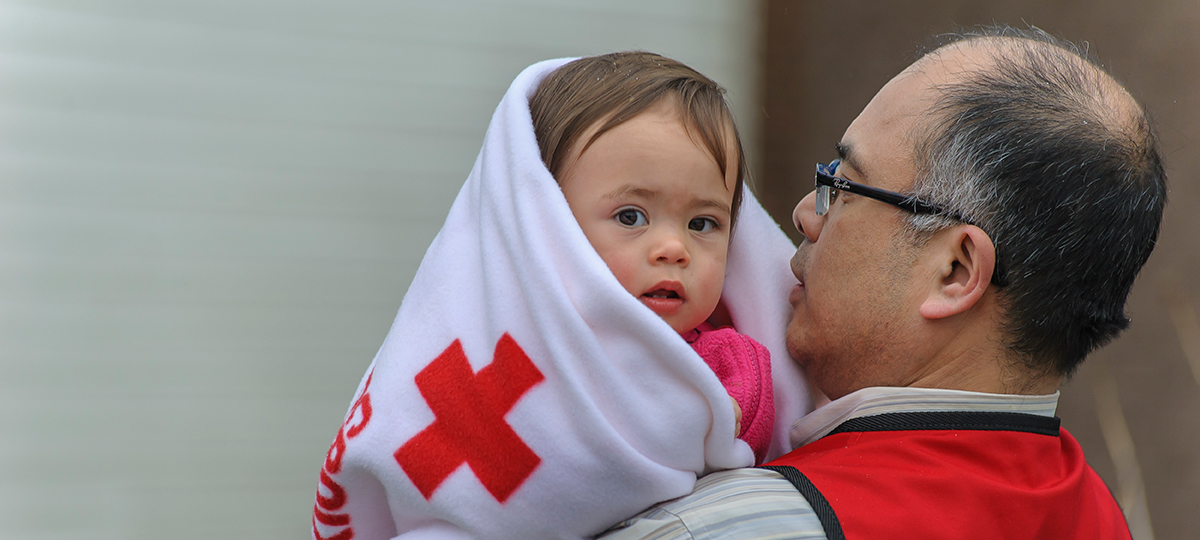 Un bénévole âgé de la Croix-Rouge apporte du réconfort à un tout-petit enveloppé dans une couverture polaire de la Croix-Rouge.