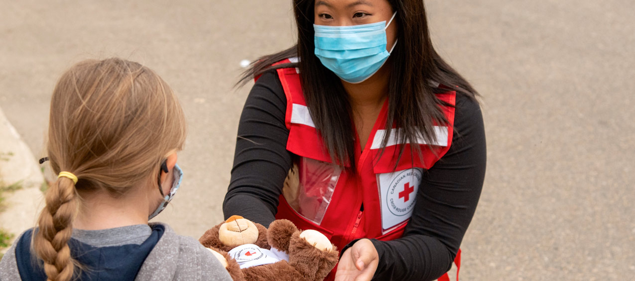 Un volontaire de la Croix-Rouge portant un masque facial tend un ours en peluche à une jeune fille.