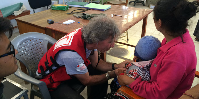 Un représentant de la Croix-Rouge portant un gilet rouge utilise un stéthoscope sur un bébé, tenu sur les genoux de sa mère.