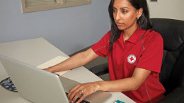 Une femme portant un polo rouge est assise à un bureau, travaillant sur un ordinateur portable.