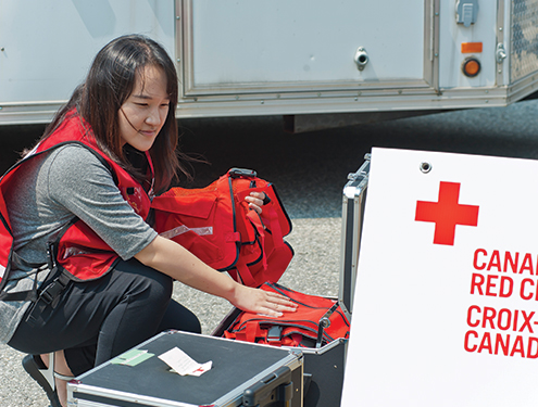 Une femme portant une veste de la Croix-Rouge est accroupie à l'extérieur, regardant dans une boîte 