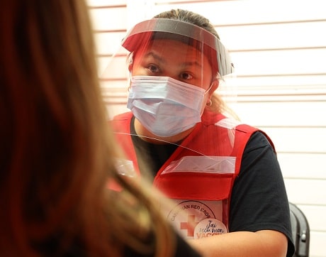 Une femme avec un masque et un écran facial, portant une veste de la Croix-Rouge, parle avec quelqu'un.