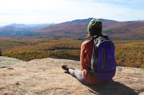 Une femme avec un sac à dos vert, assise sur un rocher, qui regarde le paysage composé de montagnes avec des arbres colorés