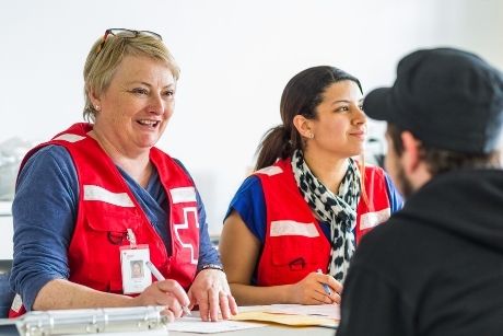 Deux personnes portant un dossard de la Croix-Rouge parlent à une personne assise devant elles. Les travailleuses ont un stylo en main et des formulaires sont sur la table.