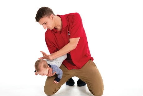 Homme avec un chandail rouge qui tient un bébé d'une main, la tête vers le bas, et qui tape entre ses omoplates de l'autre main