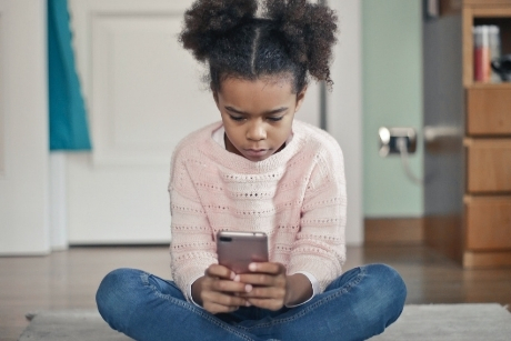 jeune fille assise en tailleur sur le sol du salon, regardant un téléphone portable dans ses mains