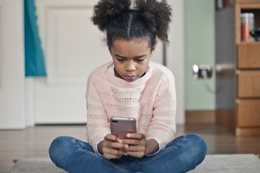 jeune fille assise en tailleur sur le sol du salon, regardant un téléphone portable dans ses mains