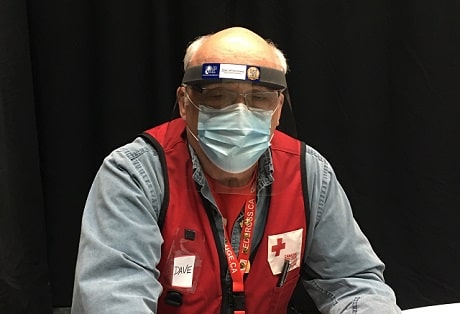An older gentleman sitting at a table in a Canadian Red Cross vest and face masks.
