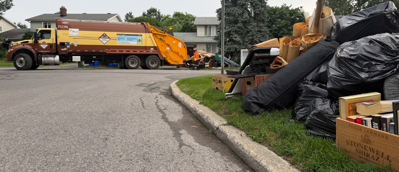 Des sacs d’ordures et des objets divers sont empilés devant les maisons dans une rue où passe un camion à ordures. 