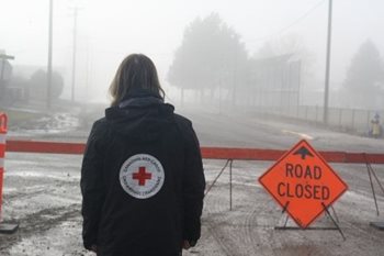 Une personne vêtue d'une veste de la Croix-Rouge observe une route fermée en raison des inondations.